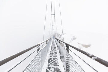 Obraz premium Hängebrücke im Schneesturm. Fußgänger überquert schmale Hängebrücke im Nebel. Suspension bridge in a snowstorm. Pedestrian crosses narrow suspension bridge in the fog.