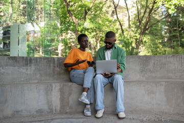 Two african american students taking break with laptop sitting near college library talking, discussing, brainstorming. Interested pleased black girl and guy studying together outdoor with computer. 