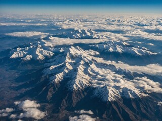 Fototapeta premium Aerial view showcases majestic mountain range covered in snow, with peaks piercing through blanket of clouds. Image captures vast expanse of rugged terrain, with snow-capped summits, deep valleys.