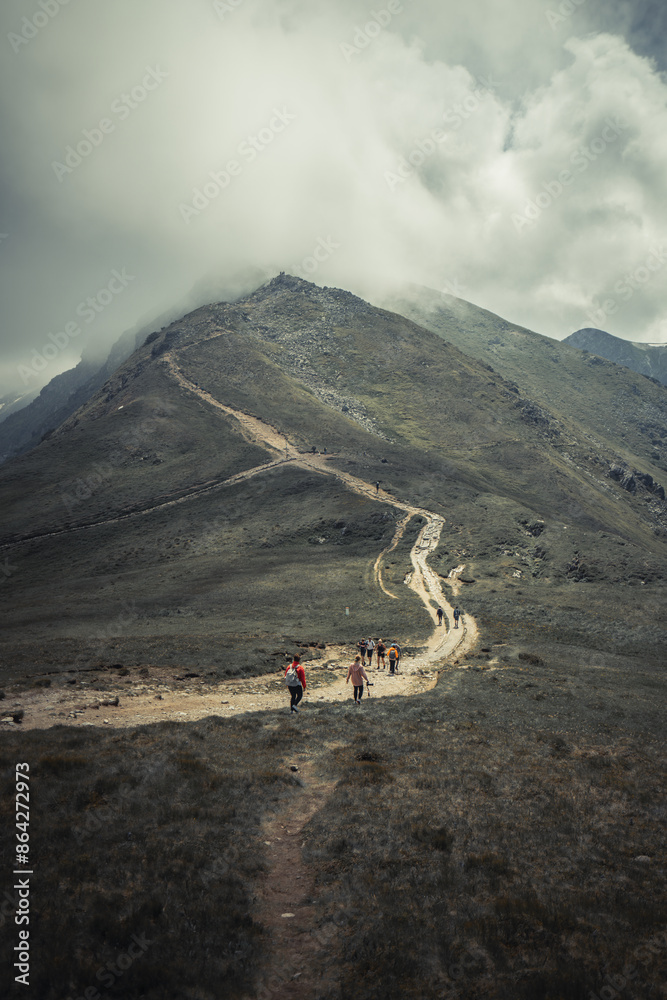 Sticker  A group walks up a dirt path beside a lush, green mountain The peak is covered with a sky filled with clouds