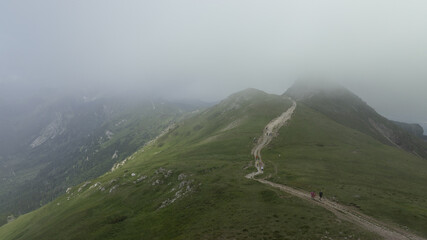  A few individuals traverse a dirt path atop a verdant hill, enshrouded in foggy conditions