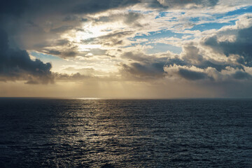 The sun shines above the ocean, piercing through fluffy clouds