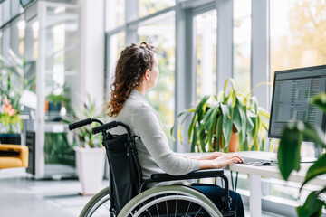 Side view of a young woman in a wheelchair using a computer in an office Concept of inclusion, diversity, accessibility and independence of people with reduced mobility.