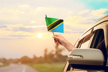 Waving the Tanzania flag against the sunrise or sunset from a car driving along a country road. Holding the Tanzania flag, traveling by car, on a weekend trip.