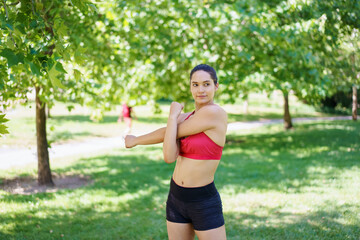 Fototapeta premium A young woman stretches in a park, wearing red sports bra and black shorts, surrounded by greenery in the sunshine