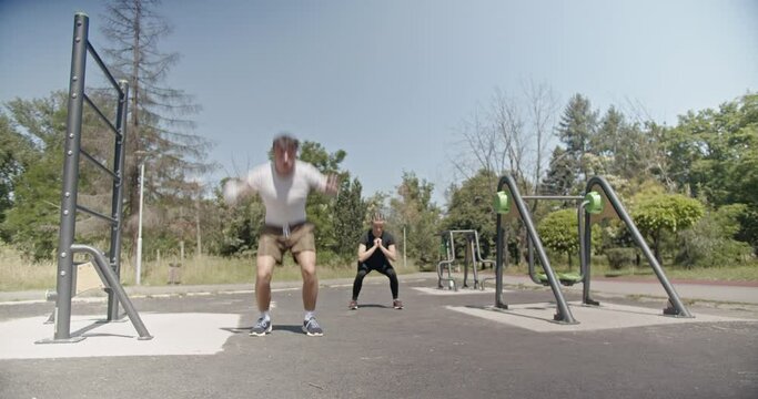 Lovely sports couple working out in a park, with a man performing exercises on parallel bars while the woman stretching.