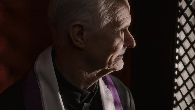 Close up shot of senior priest in traditional cassock and stole sitting inside of confessional and having conversation with penitent through lattice screen