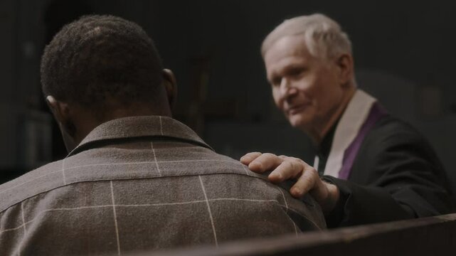 Rack focus shot of elderly priest holding hand on shoulder of young Black parishioner and giving him advice while sitting in church