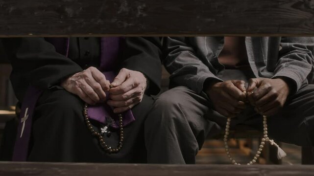 Close up shot of unrecognizable priest and parishioner sitting on bench and holding rosaries during conversation in church