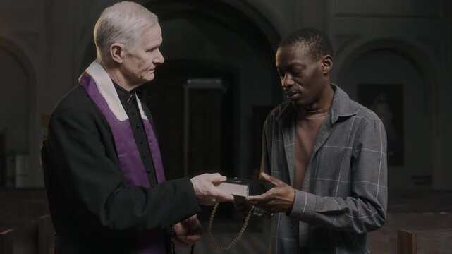 Senior Caucasian priest in traditional cassock giving Bible and talking to Black parishioner visiting church