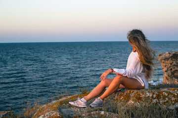 A woman with long hair sits on the rocks on the seashore at sunset. Travel and tourism