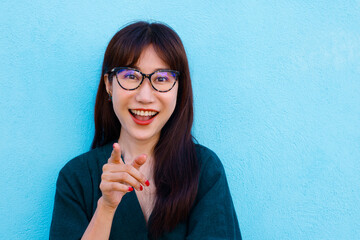Happy Asian Woman Looking And Pointing At Camera With Glasses Over Light Blue Background