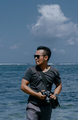 Obraz premium Asian male photographer wearing sunglasses and holding professional camera on the beach.