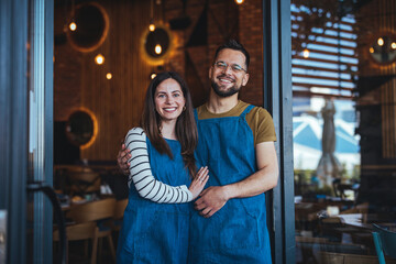 A smiling male and female barista team in aprons stand ready in a cozy coffee house, surrounded by wooden decor and brewing tools.