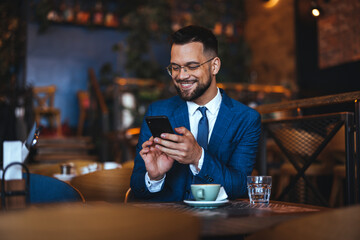 A joyful Caucasian man dressed in a formal suit interacts with his phone, enjoying a break in the cozy ambiance of a coffee house.