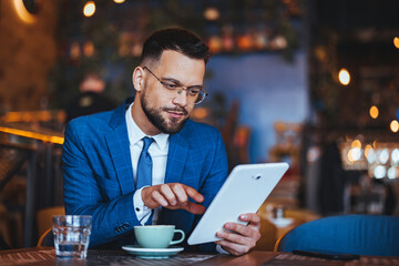 Professional Caucasian male in a sharp blue suit uses a digital tablet, managing tasks while enjoying a coffee break indoors.