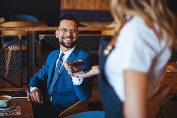 A smiling Caucasian businessman, dressed in a formal suit, makes a mobile payment to a waitress in a cozy cafeteria setting.