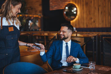 A cheerful male executive in a suit finalizes a payment using a credit card while interacting with a female server in a modern café.