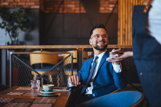 A smiling Caucasian businessman in a suit uses his smartphone for a contactless payment in a cozy cafeteria setting. His professional attire contrasts softly with the rustic interior.
