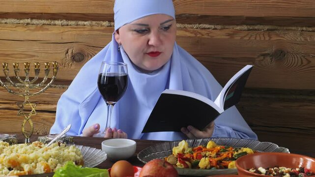 A Jewish woman in a blue veil with glasses of wine and siddur reads kiddush for wine on the Pesach Seder holiday
