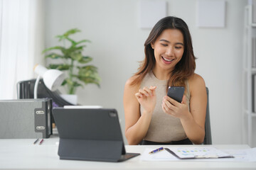 Smiling woman using smartphone at office desk with tablet, notebook, plant, and documents, showcasing a modern workspace and technology usage.