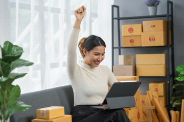 Woman celebrating success while working with a laptop from home. Surrounded by cardboard boxes, she raises her fist in excitement.