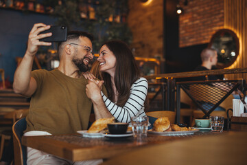 A cheerful Caucasian duo engages in taking a selfie with a smartphone in a cozy restaurant setting, seated with refreshments and pastries.