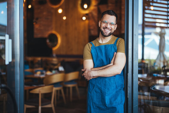 A welcoming male bartender with a genial smile stands with arms crossed, wearing a blue apron in the warm setting of a coffee house, embodying professionalism.