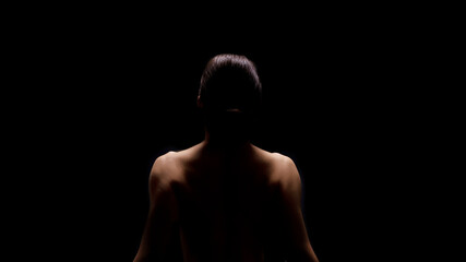 A woman sits with her back to the camera, illuminated by a single light source. She is facing away from the viewer and the background is completely black.
