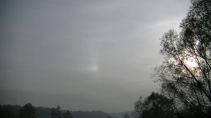 Evening grey sky with clouds Cirrus and Sundog, Mock sun, Parhelion next sun and dark trees and horizon on autumn season - timelapse. Topics: weather, meteorology, optical phenomenon, atmosphere