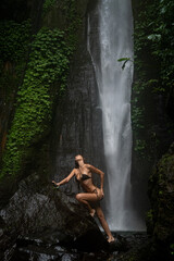 A woman in a black bikini poses on a large rock near a waterfall, arching her back, surrounded by lush tropical vegetation.