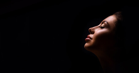 A close up shot of a womans face with eyes closed, lit from the side, with a dark background. She...