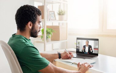 A man with a beard is seated at a desk and taking notes while listening to a woman on a video conference. He is wearing a green shirt and has a pen in his hand.