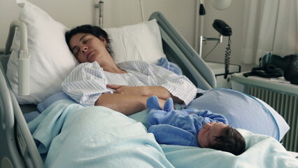 Mother and newborn baby sleeping peacefully in a hospital bed. The baby, in a blue onesie, lies on...