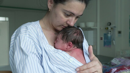 Mother holding her newborn baby close to her chest in a hospital room providing warmth and comfort. newborn wrapped in a striped shirt is peacefully asleep showcasing the bond between mother and child