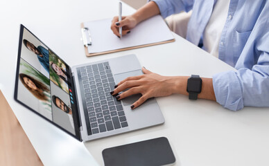 A woman manager sits at a desk, participating in a video conference on her laptop while taking notes on a clipboard. She is wearing a blue button-down shirt and a black smartwatch.