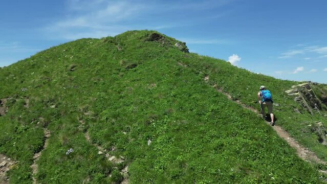 a man with a hiking backpack and trekking poles walks along the top of a picturesque mountain in green grass surrounded by clouds and high mountains with snow