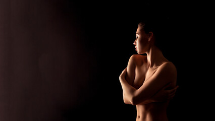 This image shows a woman standing in a dark room with her arms crossed over her chest. The lighting is soft and highlights her body and face in silhouette.