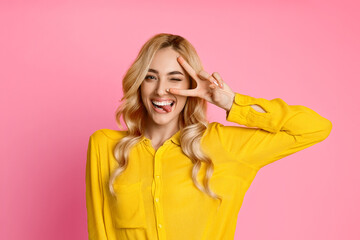 A young woman with long, blonde hair is wearing a yellow shirt and is standing against a pink background. She winks at the camera while sticking her tongue out and making a peace sign © Prostock-studio
