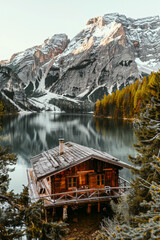 Fototapeta premium Wooden cabin nestled by a serene lake, surrounded by tall trees and dramatic snow-capped mountains, offering a breathtaking view of nature's pristine beauty.