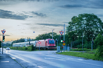 Evening passenger train with red locomotive and coaches is driving over the level grade crossing on urban setting in Ljubljana, Slovenia.