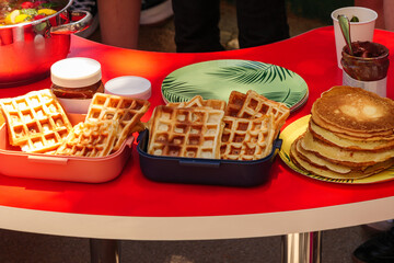 A table set for a picnic, with containers of waffles, a stack of pancakes, a chocolate spread jar, and other picnic utensils.