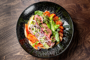 view of a gourmet dish with sliced beef, fresh herbs, tomatoes, onions, and a drizzle of sauce on a black plate.