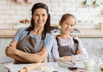 A mother and her young daughter are wearing aprons and smiling at the camera while baking in their kitchen. There are baking supplies on the counter next to them