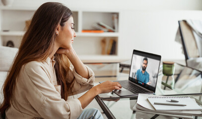 A young woman sits at a desk in her home office, attentively listening to a video conference on her laptop. The room is well-lit, and the woman is dressed casually in a light-colored shirt.