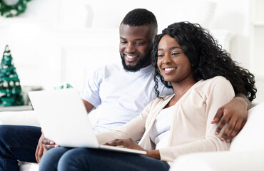 A couple sits together on a white couch, smiling as they browse a laptop computer.