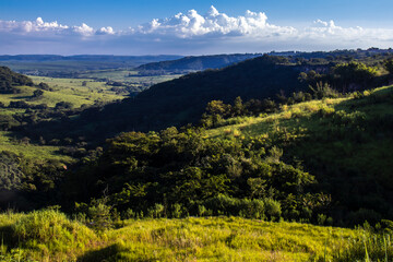 Colorful view of sunlit green mountain valley with forest, river, eucalyptus trees and grassland. against mountain range under cloudy sky in Brazil. Wide mountain valley in sunlight.