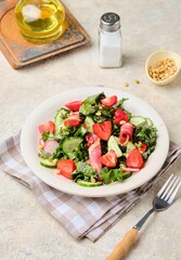 A healthy salad with cabbage kale, fresh cucumbers, radish, strawberries and smoked meat on a beige ceramic plate on a light concrete background.