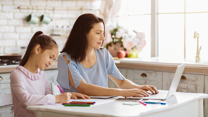 Obraz premium A woman sits at a table in a kitchen, working on a laptop computer while her daughter colors at the table beside her.