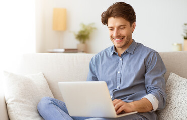 A man sits on a couch in his home, smiling as he uses a laptop.